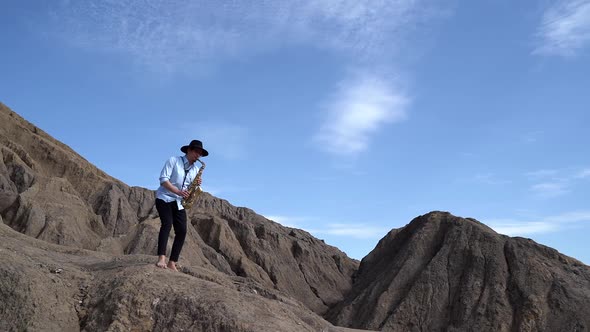 A Musician in a Hat Plays Jazz on a Saxophone Standing on Mountains and Rocks on a Warm Sunny Day.