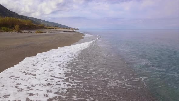 Drone flies over sea coast, aerial view of waves reaching shore and splashing mountains on horizon