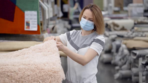 A Young Woman Wearing a Protective Mask at a Home Goods Store. The Girl Chooses a Carpet in Her