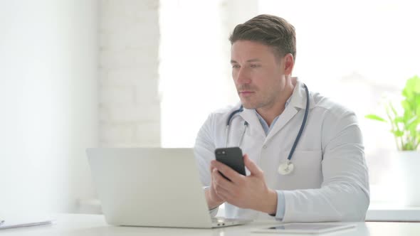 Male Doctor Browsing Internet on Smartphone while using Laptop in Office