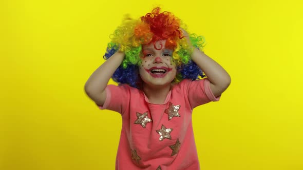 Little Child Girl Clown in Colorful Wig Making Silly Faces, Shouts, Grabs Her Head, Waves Her Hands