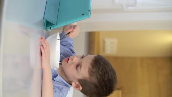 Little Boy Eating Cereal with Milk in the Kitchen and Watching Cartoon on Tablet Computer