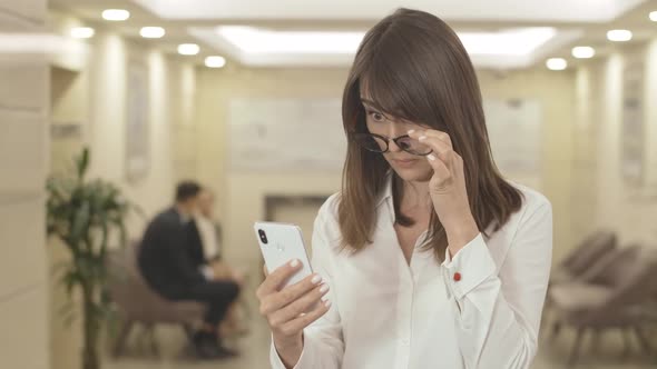 Portrait of Confused Young Brunette Woman in Eyeglasses Trying To Read on Smartphone Screen