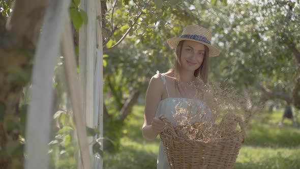 Pretty Young Woman in Straw Hat Holding the Wicker Basket with Herbs and Looking at the Camera