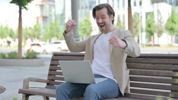 Young Man Celebrating Success on Laptop While Sitting on Bench