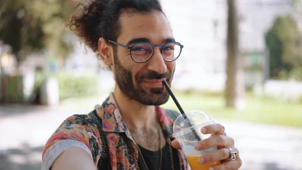 Smiling curly-haired bearded man in eyeglasses drinking juice at the camera