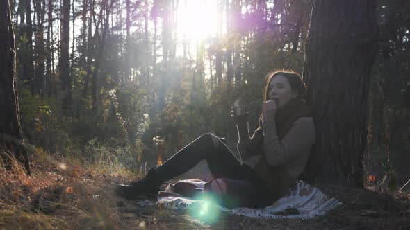 Happy young woman tourist drinking hot tea and eating cookies relaxing in autumn forest at sunset