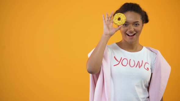 Cute Girl Having Fun Looking Through Donut, Bakery Products, Yellow Background
