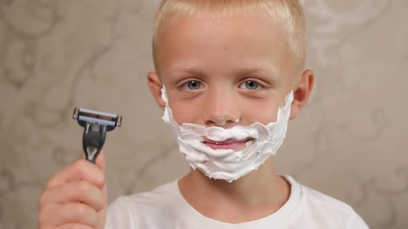 A Little Boy with Shaving Foam on His Face Holds a Shaving Machine in His Hands