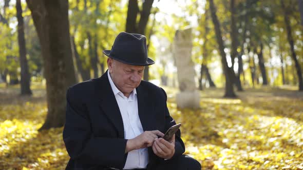 Joyful Senior Man in Suit and Hat Uses Smartphone and Talks with Smile in Park