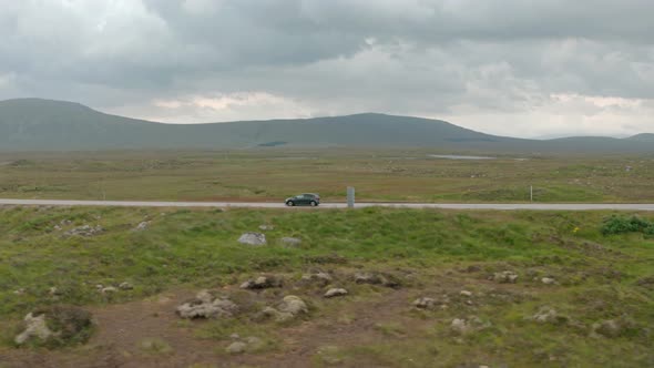 Profile follow drone shot of small black hatchback car driving through scottish highlands