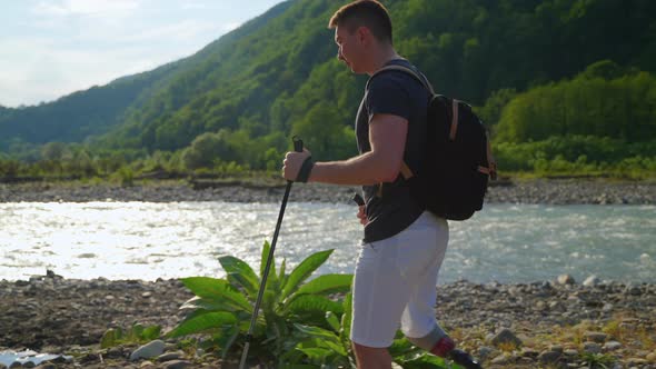 Male Backpacker with Prosthetic Leg is Strolling Over Shore of River in Summertime