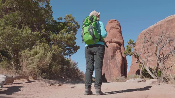 Elderly Active Female Tourist With Backpack Looks Around At Red Rocks In Desert