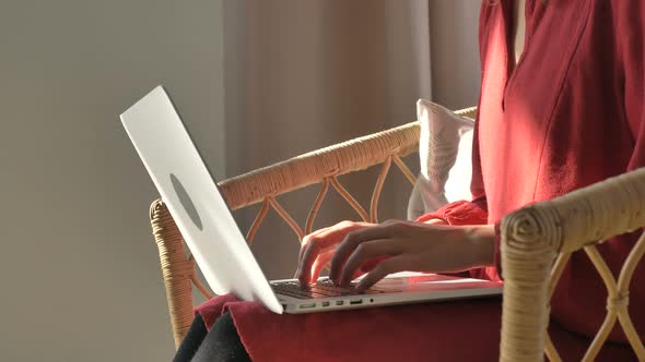 Lady in Red Dress Types on Laptop Sitting in Rattan Armchair