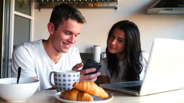 Couple using mobile phone and laptop while having breakfast