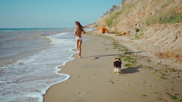 Slow Motion Rear View of Happy Joyful Fitness Girl in Bikini Running with Dog Along Sand Sea Beach