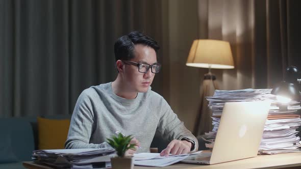 Asian Man Looking At Laptop And Taking Note While Working With Documents At Home