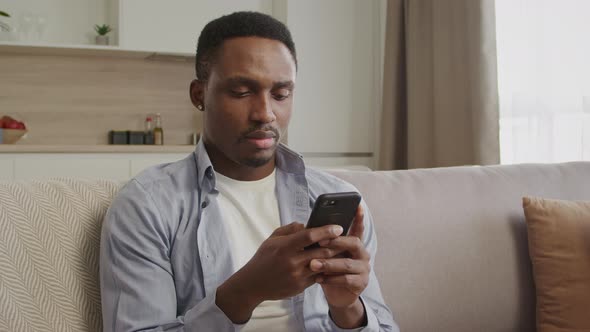 Portait of an African American Guy Thinking and Typing on a Smart Phone in His Apartment
