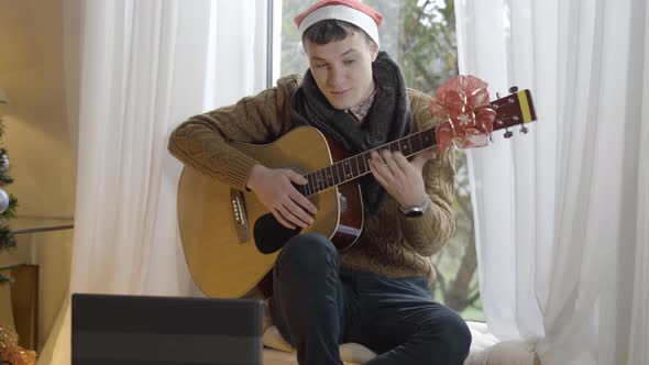 Portrait of Young Brunette Man in Christmas Hat Playing Guitar and Talking in Laptop Video Chat
