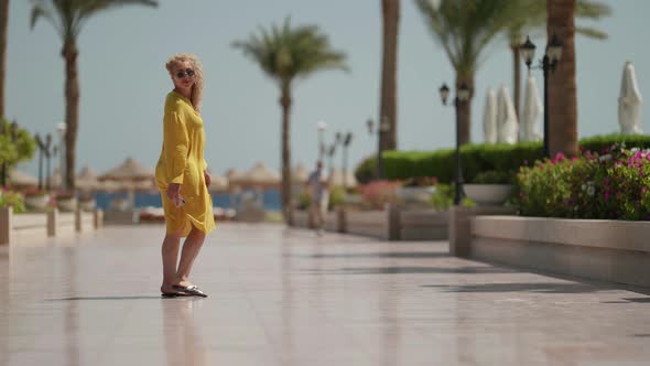 a Curly Blonde in Sunglasses and a Yellow Fluttering Dress Turns Around and Walks Down the Alley to