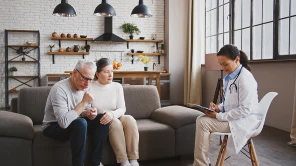 Coughing Senior Man and His Wife Talking to Female Doctor About Complaints and Symptoms of Disease