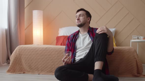 Depressed man is sitting on floor with glass of alcohol in hand with an absent look, front view