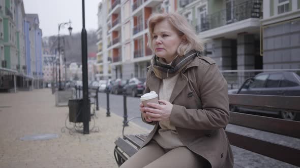 Sad Senior Caucasian Woman Sitting on Bench Outdoors and Drinking Coffee. Lonely Elegant Retiree