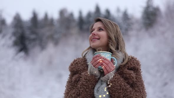 Beautiful girl in coat with cup of hot drink in a snow forest.