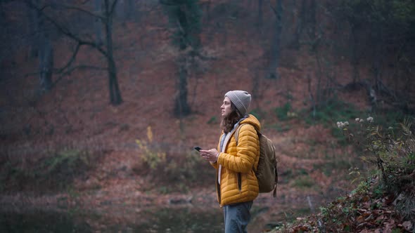 A Young Woman with a Backpack Using Her Phone While Standing at the Forest Lake