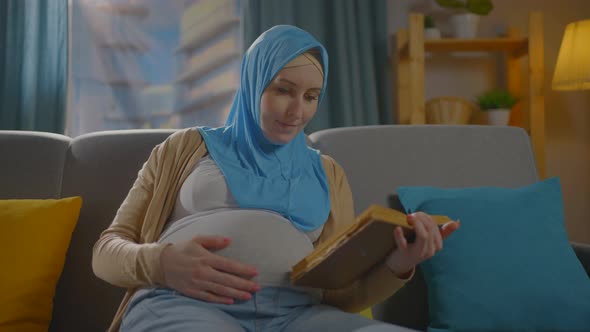 Pregnant Young Muslim Woman in a Hijab Scarf Sitting on the Sofa Reading the Book of the Koran