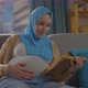 Pregnant Young Muslim Woman in a Hijab Scarf Sitting on the Sofa Reading the Book of the Koran - VideoHive Item for Sale