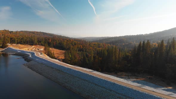 Aerial shot of a dam on a blue alpine lake surrounded by pine trees and mountains