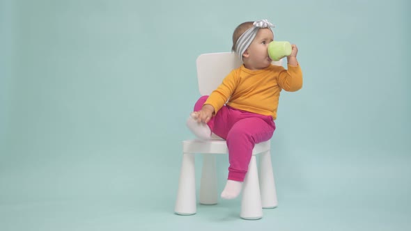 Toddler 12-17 months old sits on a small white chair and drinks yogurt from a baby cup