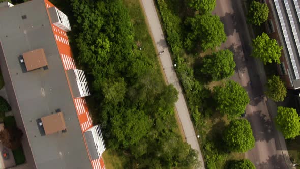 City street, sidewalk, greenery and apartment building rooftops with parking lot, aerial top down vi