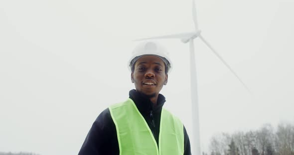 A Man in Work Uniform Smiles Looking at the Camera Standing Near a Wind Turbine