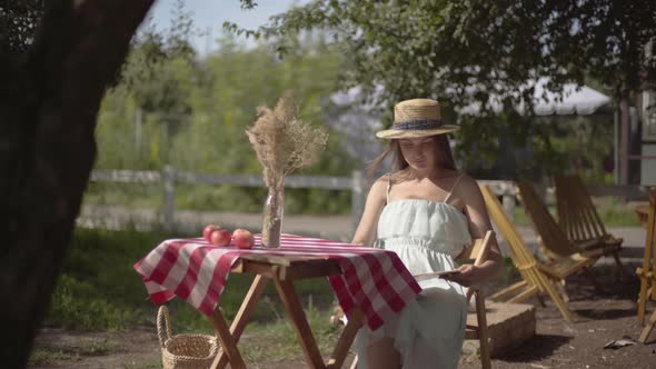 Young Rural Girl in a Straw Hat and White Dress Sitting at the Small Table with Vase with Decorative