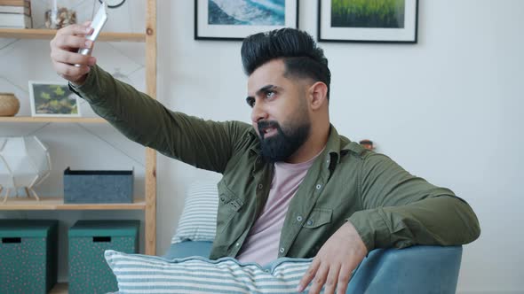 Young Middle Eastern Man Taking Selfie Using Smartphone Posing for Camera with Vsign at Home