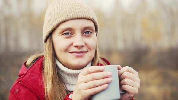 Woman is Sitting in Autumn Forest Drinking Hot Tea From Gray Mug and Enjoying Nature