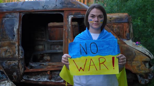Portrait of Young Sad Woman with No War Placard Standing at Car Dump with Burnt Down Automobiles