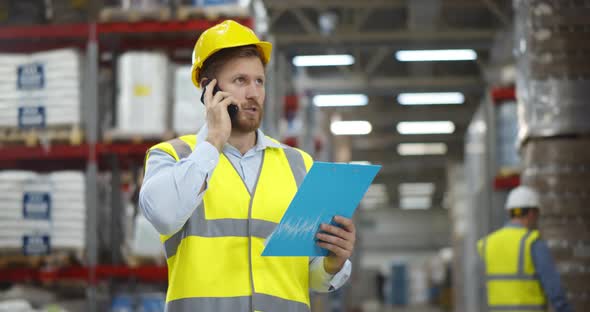 Male Worker Looking at Clipboard While Talking on Mobile Phone in Warehouse