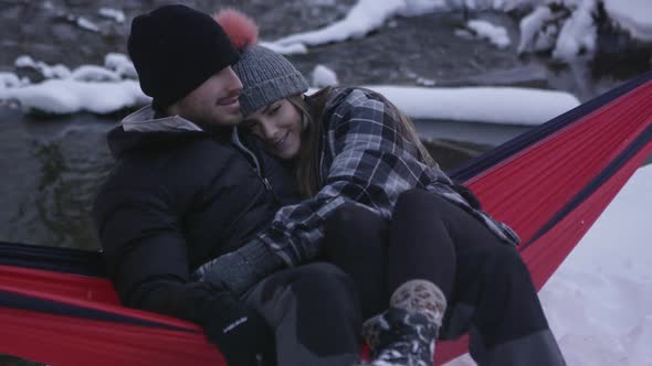 Young man and woman rocking in hammock as they cuddle outside in winter