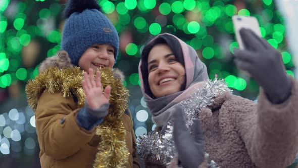 Positive Woman and Boy Waving at Selfie Camera on Smartphone Live Streaming with Christmas Lights at