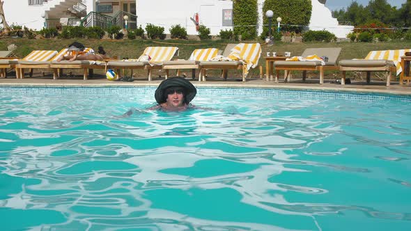 Caucasian Senior Female Elderly in Black Hat Swims in the Blue Water Pool in Hotel
