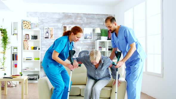 Female Doctor and Her Assistant Helping Old Woman with Crutches To Stand Up From the Couch