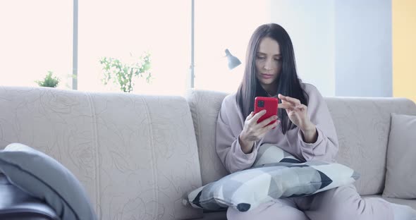 Sad Woman Sits on Sofa and Looks Into Smartphone