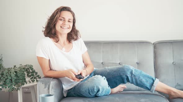 Young Woman Sitting on a Sofa with a TV Remote Control and Switching Channels.