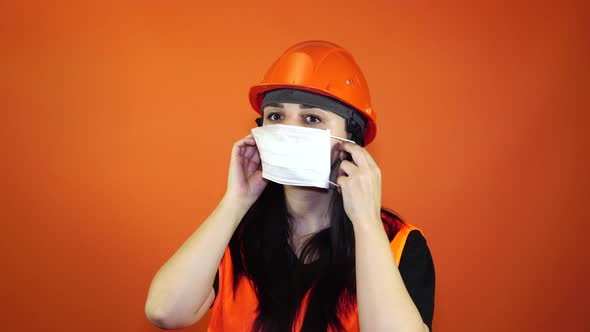 Female Construction Worker in Overalls Putting on Medical Mask on Face on Orange Background