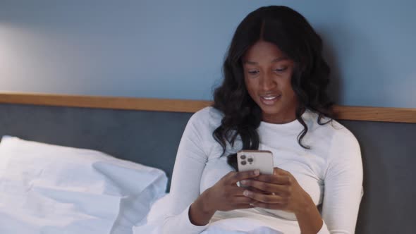Smiling African woman typing by phone in the bed