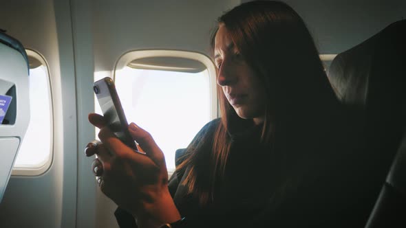 Woman Sitting Near Airplane Window and Using Mobile Phone During Flight.