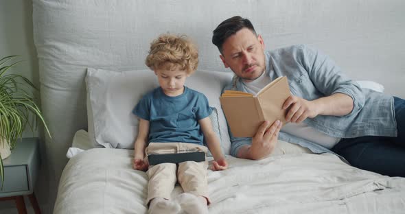 Serious Guy Reading Book in Bed While Small Child Watching Cartoons on Tablet